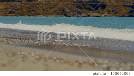 Waves crash on the shore at a beach during the midday sun with clear water and pebbles scattered on the sand Waves crash on the shore at a beach during the midday sun with clear water and pebbles scattered on the sand 138651063