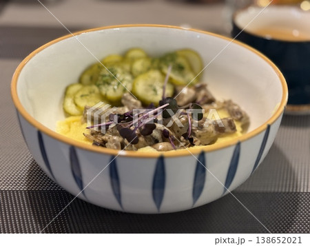 Ceramic bowl with beef stroganoff, mashed potatoes, and sliced cucumbers on table 138652021