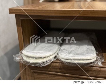 Two pairs of cozy beige slippers wrapped in plastic placed on wooden shelf in bedroom setting 138652028
