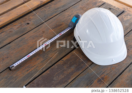 White construction hard hat and tape measure on a wooden background. Safety helmet and measuring tool on rustic timber surface. Building and renovation concept. White construction hard hat and tape measure on a wooden background. Safety helmet and measuring tool on rustic timber surface. Building and renovation concept. 138652528