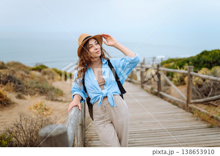 A happy young woman stands on a wooden boardwalk near the beach, wearing a straw hat and blue shirt 138653910