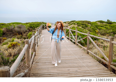 Young woman with curly hair wears a stylish hat and waves while taking a selfie. Travel, tourism 138653953