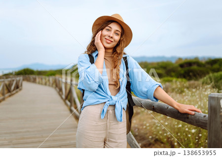 A happy young woman stands on a wooden boardwalk near the beach, wearing a straw hat and blue shirt A happy young woman stands on a wooden boardwalk near the beach, wearing a straw hat and blue shirt 138653955