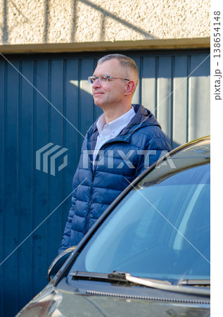 Adult man aged 51 wearing glasses poses near car with building background Adult man aged 51 wearing glasses poses near car with building background 138654148