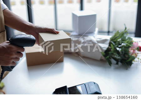 A person using a barcode scanner to scan a small cardboard box in a retail shop for inventory management or preparing an order for shipping. A person using a barcode scanner to scan a small cardboard box in a retail shop for inventory management or preparing an order for shipping. 138654160