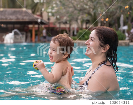 Caucasian woman holds son while swimming in pool on vacation. Caucasian woman holds son while swimming in pool on vacation. 138654311