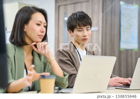 フリーアドレスのオフィスで女性の先輩に相談しながら作業する若い男性社員 フリーアドレスのオフィスで女性の先輩に相談しながら作業する若い男性社員 138655646