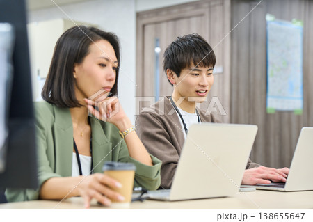 フリーアドレスのオフィスで女性の先輩に相談しながら作業する若い男性社員 138655647