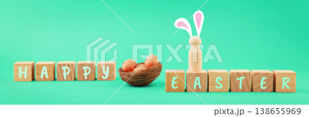 Minimalist Happy Easter banner featuring wooden blocks, a nest with speckled eggs, and a cute wooden peg doll with bunny ears on a vibrant green background. High quality photo 138655969