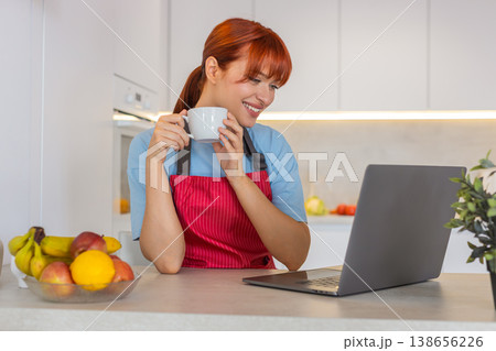 Young woman in kitchen with laptop answers emails writes replies and drinks coffee focused workflow 138656226