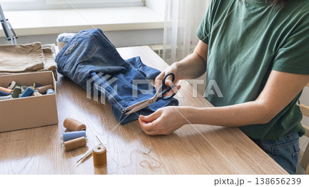Hands cutting thread from old jeans for reuse, promoting sustainable fashion and circular economy principles. Person upcycling denim jeans with scissors, repairing clothing 138656239