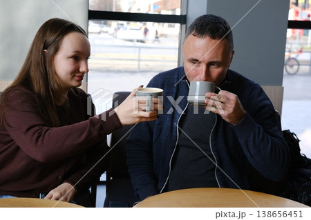 European father in blue sweater and 17-year-old daughter in brown sweater enjoy tea at a cafe. Cozy family moment in city. European father in blue sweater and 17-year-old daughter in brown sweater enjoy tea at a cafe. Cozy family moment in city. 138656451
