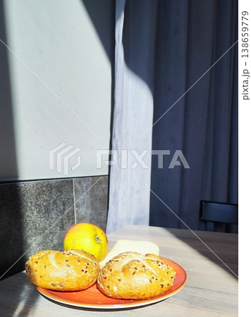 A sunlit wooden table holds a red plate with two round Kaiser bread rolls topped with seeds and an apple  138659779