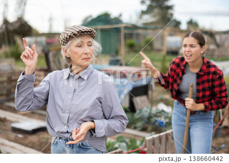 Two upset young and old female neighbors disagree with each other while digging garden on smallholding in autumn Two upset young and old female neighbors disagree with each other while digging garden on smallholding in autumn 138660442