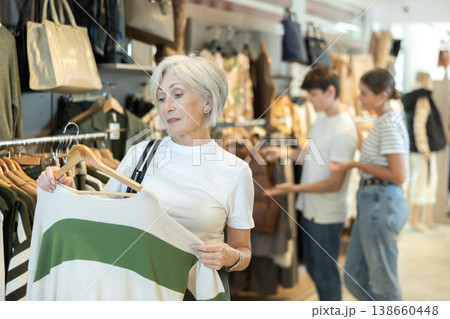 Senior woman customer in store has removed hanger with jersey from window and is considering product Senior woman customer in store has removed hanger with jersey from window and is considering product 138660448