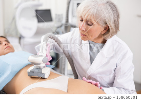 Young woman getting ultrasound cavitation body contouring treatment, anti-cellulite therapy, vacuum massage from old female cosmetologist. Hardware cosmetology for body sculpting Young woman getting ultrasound cavitation body contouring treatment, anti-cellulite therapy, vacuum massage from old female cosmetologist. Hardware cosmetology for body sculpting 138660501