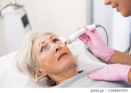 Young beautician doctor doing RF lifting procedure for face lifting of mature woman laying in beauty salon. Hardware cosmetology Young beautician doctor doing RF lifting procedure for face lifting of mature woman laying in beauty salon. Hardware cosmetology 138660703