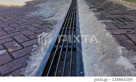 Metal storm drain grate on brick paved walkway. A long metal drainage grate runs through a brick-paved walkway and concrete gutter. The perspective shows parallel steel bars for water management in an Metal storm drain grate on brick paved walkway. A long metal drainage grate runs through a brick-paved walkway and concrete gutter. The perspective shows parallel steel bars for water management in an 138663200