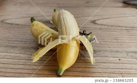 Peeled ladyfinger banana on wooden table close up. A cluster of small ripe ladyfinger bananas on a rustic wooden background with one banana partially peeled and standing upright in the foreground. Peeled ladyfinger banana on wooden table close up. A cluster of small ripe ladyfinger bananas on a rustic wooden background with one banana partially peeled and standing upright in the foreground. 138663205