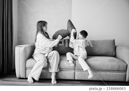 Two young siblings started a pillow fight on couch. Little boy and her older sister fighting by pillows at home. Funny spending time with family. Black and white image. Two young siblings started a pillow fight on couch. Little boy and her older sister fighting by pillows at home. Funny spending time with family. Black and white image. 138666063