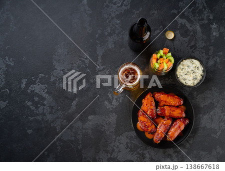 bbq buffalo chicken wings in a plate, with beer, carrot and celery sticks on a dark table, top view. 138667618