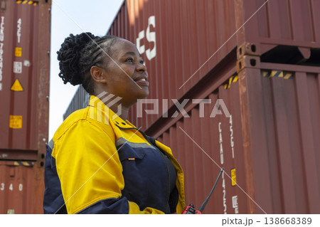 Black foreman woman worker talk on walkie-talkie at container yard, holding laptop computer. Logistics import export shipping concept. Black foreman woman worker talk on walkie-talkie at container yard, holding laptop computer. Logistics import export shipping concept. 138668389