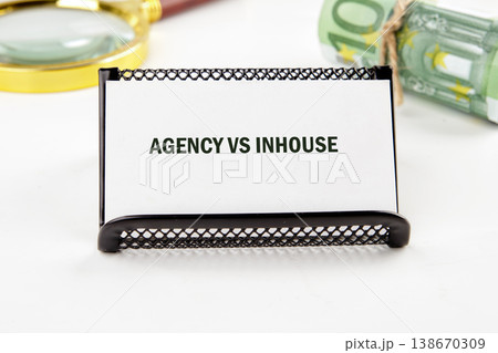 A white card on a black stand shows AGENCY VS INHOUSE. A magnifying glass and rolled Euro banknotes sit nearby A white card on a black stand shows AGENCY VS INHOUSE. A magnifying glass and rolled Euro banknotes sit nearby 138670309