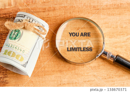 A magnifying glass reveals the phrase You Are Limitless. A bundle of US hundred Dollar bills tied with twine rests on a warm wood surface nearby A magnifying glass reveals the phrase You Are Limitless. A bundle of US hundred Dollar bills tied with twine rests on a warm wood surface nearby 138670371