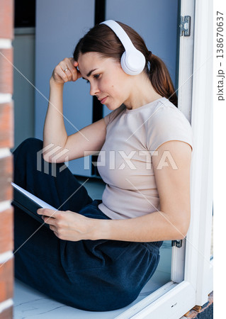 Young woman is sitting by the window wearing headphones and thoughtfully looking at her notebook. Student. Remote work. 138670637
