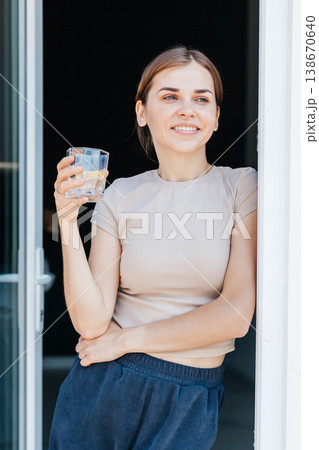 A happy young woman with a smile on her face enjoys a refreshing drink, standing in the doorway and bathed in natural light. 138670640