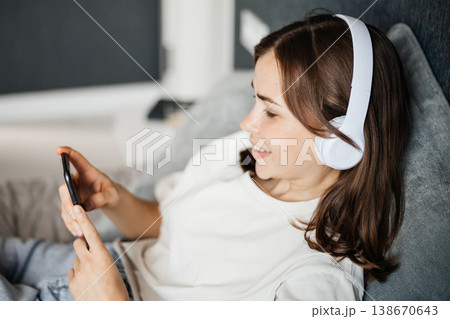 Young woman wearing headphones and looking at her smartphone while relaxing on a couch 138670643