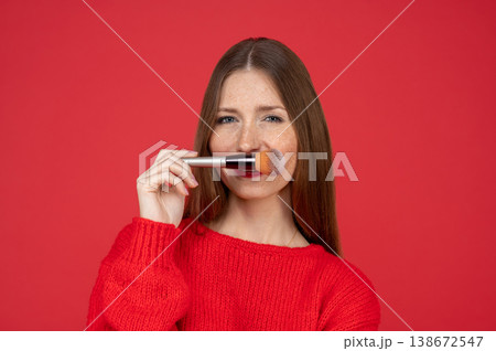 Happy young woman with freckles wearing red sweater applying powder with makeup brush 138672547