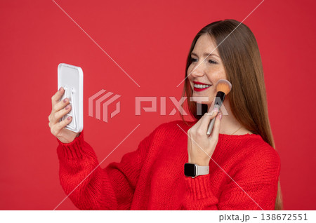 Cheerful young woman with freckles applying makeup with brush while looking in small mirror 138672551