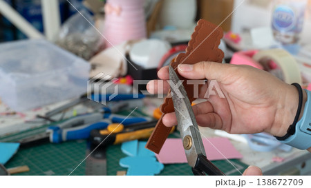 Craftsman hand holds a sheet of brown leather. Using old black handled scissors to cut. Blurred background miscellaneous element the craft. Both the cut pieces of leather and the various work tools. Craftsman hand holds a sheet of brown leather. Using old black handled scissors to cut. Blurred background miscellaneous element the craft. Both the cut pieces of leather and the various work tools. 138672709