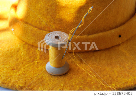 Close up of a yellow thread spool with a sewing needle and loose thread resting on a yellow felt hat brim. Close up of a yellow thread spool with a sewing needle and loose thread resting on a yellow felt hat brim. 138673403