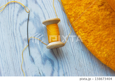 Top view of a yellow thread spool and sewing needle with a loose thread and a felt hat edge on a wooden table Top view of a yellow thread spool and sewing needle with a loose thread and a felt hat edge on a wooden table 138673404
