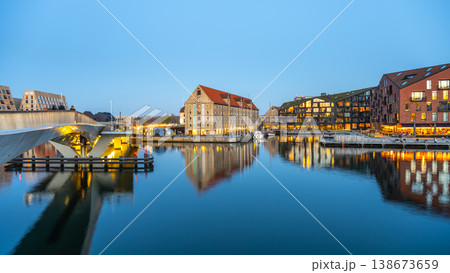 People walk along the waterfront at Kroyers Plads in Christianshavn, Copenhagen. Boats are docked and lights reflect on the calm water as evening falls. 138673659