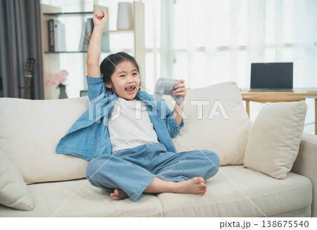 Joyful Girl Celebrating Victory on Couch While Playing Video Game, Wearing Casual Blue Outfit with Excitement and Happiness in Modern Living Room Setting Joyful Girl Celebrating Victory on Couch While Playing Video Game, Wearing Casual Blue Outfit with Excitement and Happiness in Modern Living Room Setting 138675540