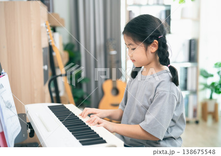 Young Girl Playing Piano in Bright Room with Guitar and Plants, Focused on Music Practice, Nurturing Creativity and Talent in Home Environment Young Girl Playing Piano in Bright Room with Guitar and Plants, Focused on Music Practice, Nurturing Creativity and Talent in Home Environment 138675548