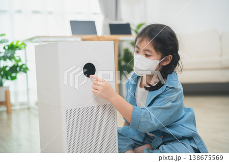 Young girl in a face mask interacts with a modern air purifier in a bright living room setting, promoting health and safety at home for children. 138675569