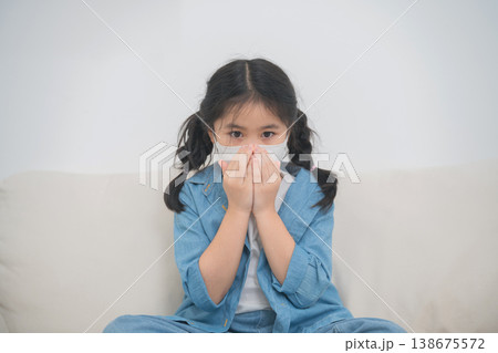 Young girl wearing a protective mask sitting on a couch at home, demonstrating caution and concern during health crisis or pandemic situation 138675572