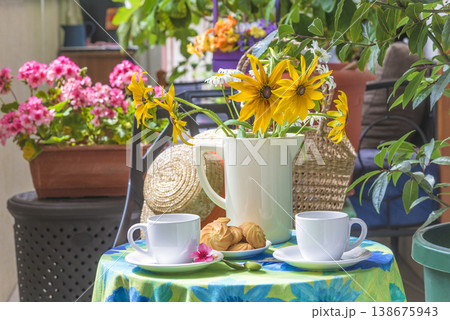 Summer still life: two white cup of coffee, cookies, yellow flowers in vase on a table covered by tableclothe on a terrace Summer still life: two white cup of coffee, cookies, yellow flowers in vase on a table covered by tableclothe on a terrace 138675943
