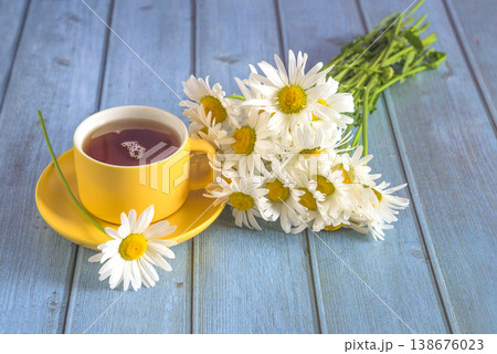 Yellow cup of tea and bouquet of white daisies flowers on blue wooden background; close up Yellow cup of tea and bouquet of white daisies flowers on blue wooden background; close up 138676023