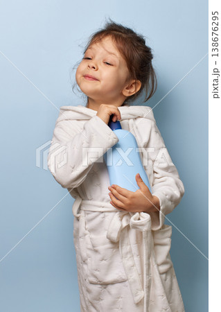 Young child girl in bathrobe closing eyes, happy holding a blue lotion bottle 138676295