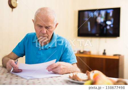 Serious older man sitting at table writing letter or filling some paper form Serious older man sitting at table writing letter or filling some paper form 138677498