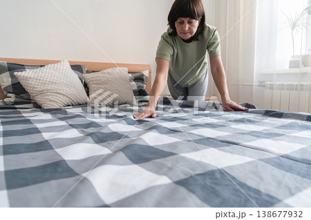 An adult woman tidying a bedroom, arranging a duvet cover with a checkered pattern and pillows, performing daily domestic chores. Authentic lifestyle 138677932
