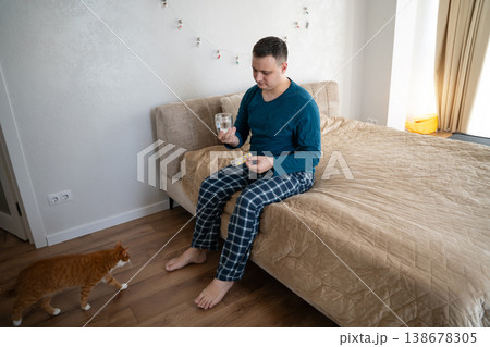 A 35 year old caucasian man sits on his bed in his bedroom with pills and water in his hands 138678305