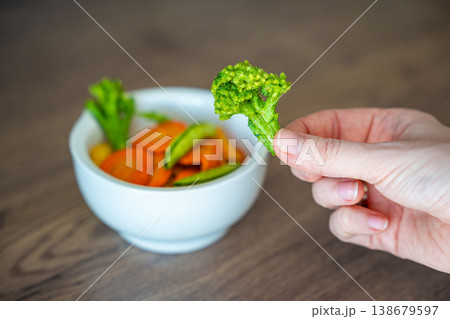 Hand holding broccoli vegetable chip above bowl on wooden table. Healthy snack alternative and clean eating lifestyle concept. Hand holding broccoli vegetable chip above bowl on wooden table. Healthy snack alternative and clean eating lifestyle concept. 138679597