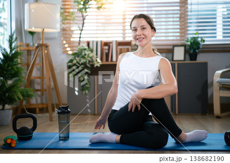 Smiling woman sitting on yoga mat after indoor exercise session Smiling woman sitting on yoga mat after indoor exercise session 138682190