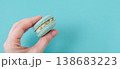Female hand holding single turquoise macaron to show texture, mint flavored french cookie against blue background 138683223